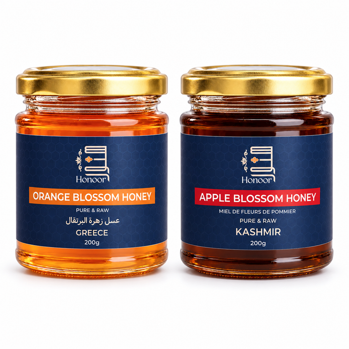 Two glass jars of Honoor brand honey on a white background: Orange Blossom Honey from Greece on the left and Apple Blossom Honey from Kashmir on the right. Both jars have gold lids and dark blue labels with orange and red text.