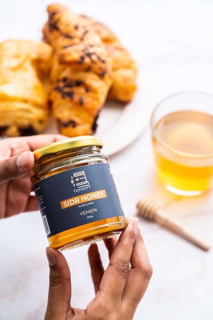 Hand holding a jar of Sidr Honey with pastries and a glass of honey in the background.

