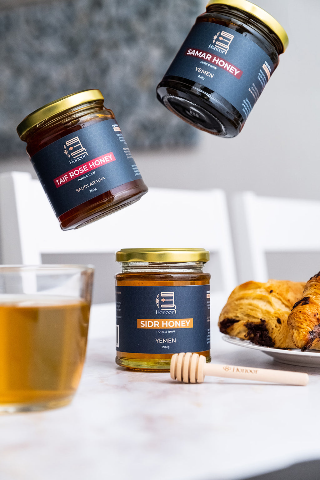 Three jars of Samar Honey with a glass of honey and a plate of food on a table.

