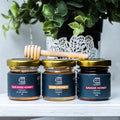 Three jars of honey with a honey dipper on a white surface with greenery in the background

