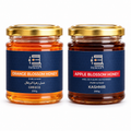 Two glass jars of Honoor brand honey on a white background: Orange Blossom Honey from Greece on the left and Apple Blossom Honey from Kashmir on the right. Both jars have gold lids and dark blue labels with orange and red text.
