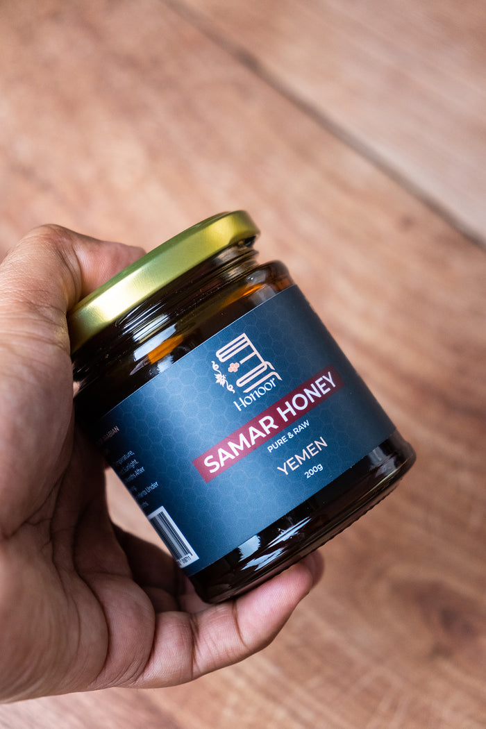 Hand holding a jar of Samar Honey with a blue label on a wooden surface

