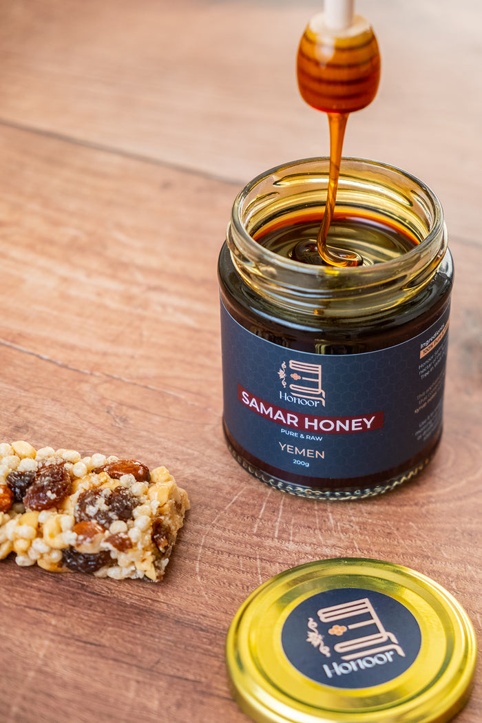 Jar of Samar Honey with honey being drizzled over a piece of food on a wooden surface.

