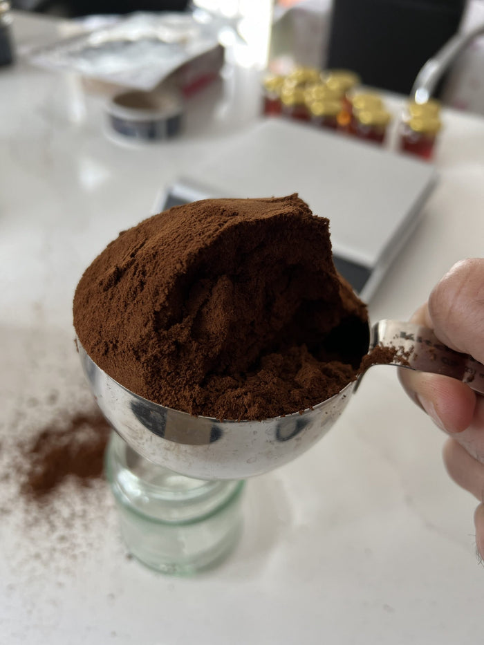Cocoa powder in a metal scoop held over a glass container on a kitchen counter.

